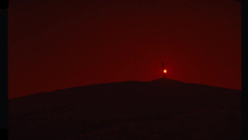 A person holding a flashing light stands on a distant hill at dusk under a deep red sky