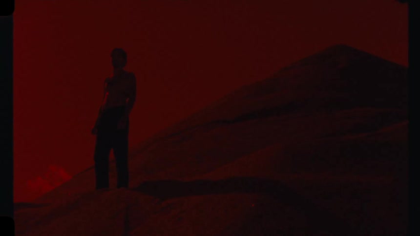 A mysterious shirtless man stands on a sandy hill in a desolate landscape under a deep red sky