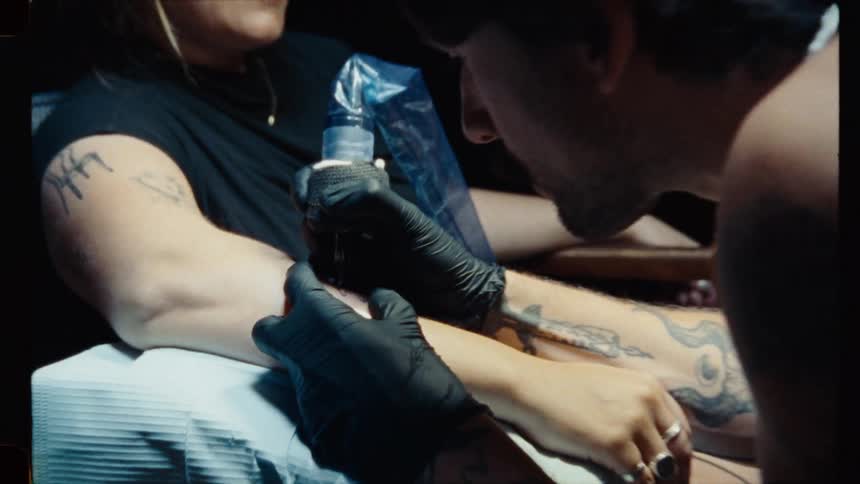 A tattoo artist carefully inks a design on a woman's arm in a dimly lit studio, captured with a vintage film aesthetic