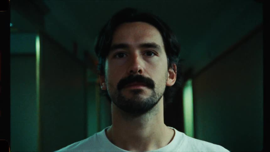 A slow zoom-out reveals a contemplative man with a mustache standing in a moody, green-lit hallway with reflective walls