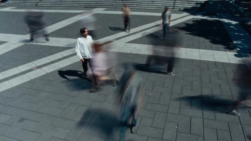Slow-shutter footage from a high angle shows a man walking as a blurred crowd moves around him in a city square