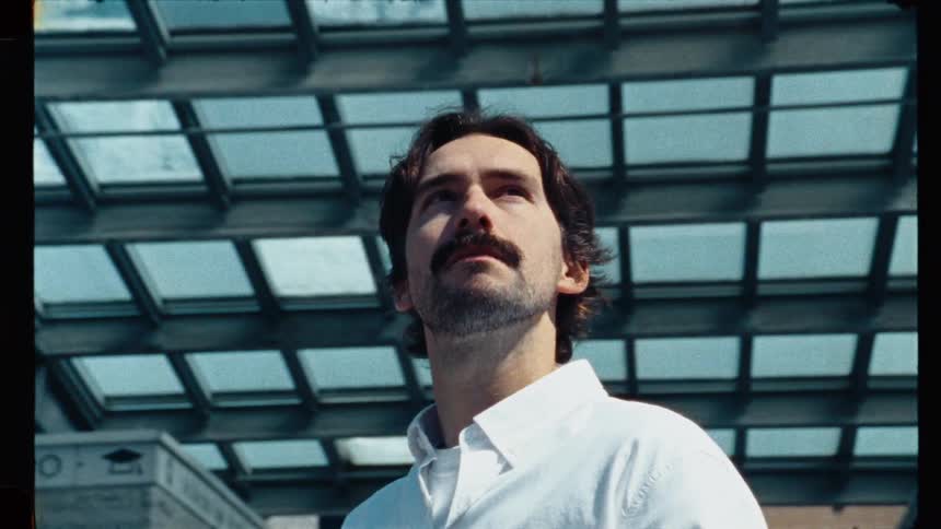Low-angle vintage film shot of a thoughtful man with a mustache looking up under a modern glass and steel canopy