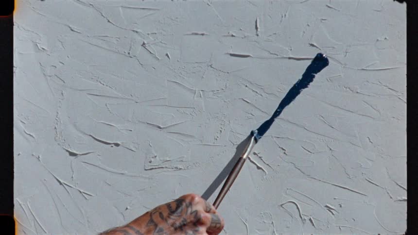 A tattooed artist slowly drags a paintbrush with blue paint across a blank, textured canvas, creating a single abstract stroke