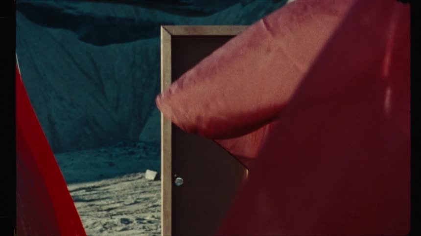 A surreal, vintage film-style shot of a lone door standing in a desolate landscape as red fabric blows in the foreground