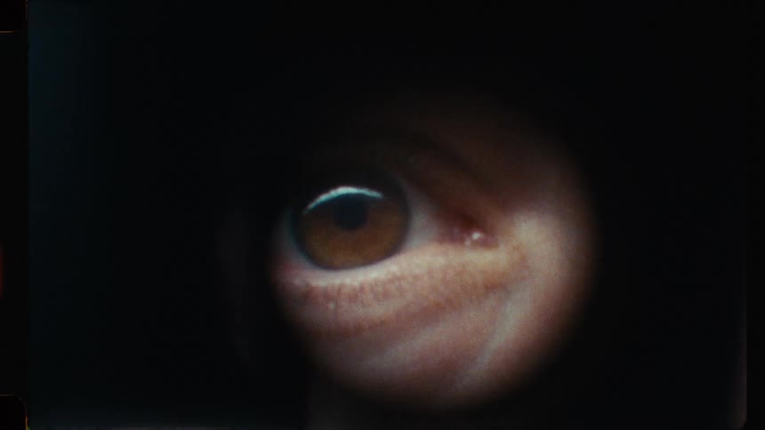A man with a mustache approaches a door and peers through the peephole in a suspenseful, vintage film-style close-up of his eye