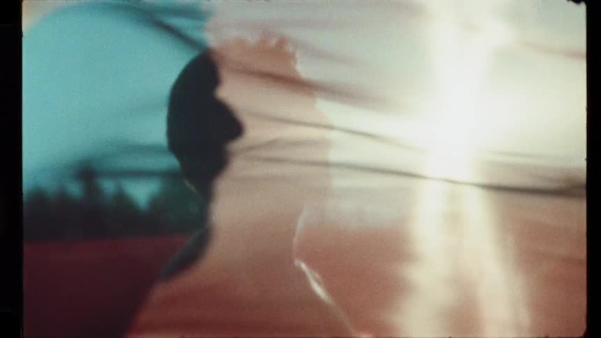 Vintage-style footage shows a silhouetted man with tattoos enjoying the bright sun as red fabric blows in the wind