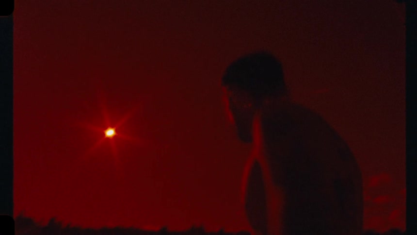 A silhouetted tattooed man dances against a surreal, deep red sky at dusk in this grainy, vintage-style film footage