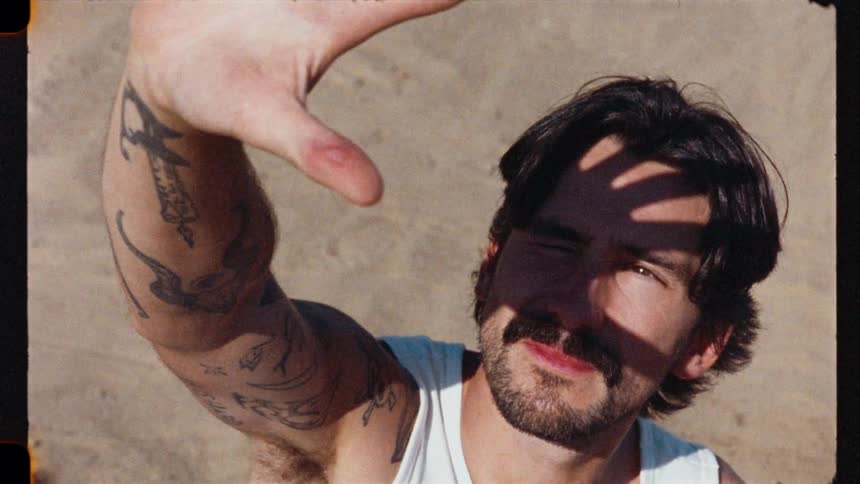 High-angle, vintage-style shot of a tattooed man with a mustache shielding his eyes from the bright sun