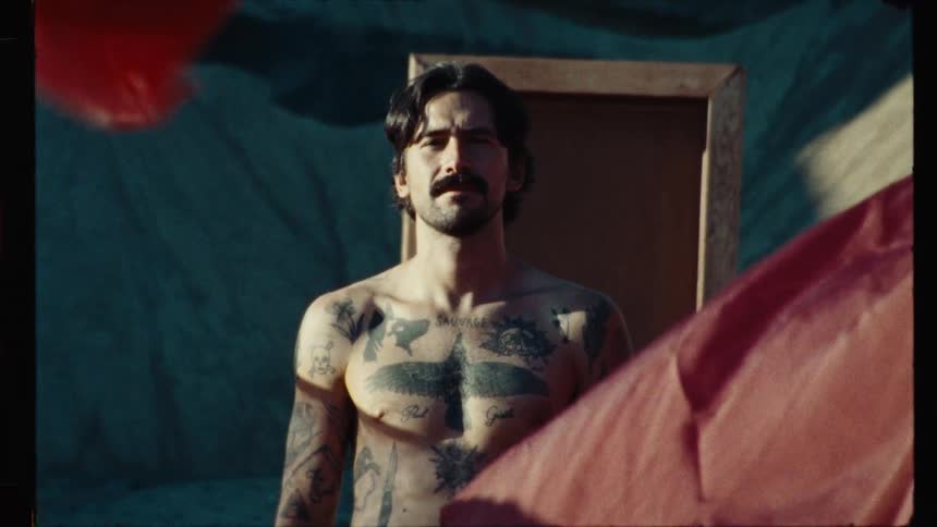 A vintage film shot captures a shirtless man with tattoos and a mustache looking at the camera as red fabric waves in the foreground