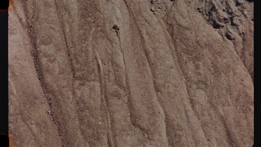 A vintage, static, close-up shot of sand with visible erosion lines on a sloped surface