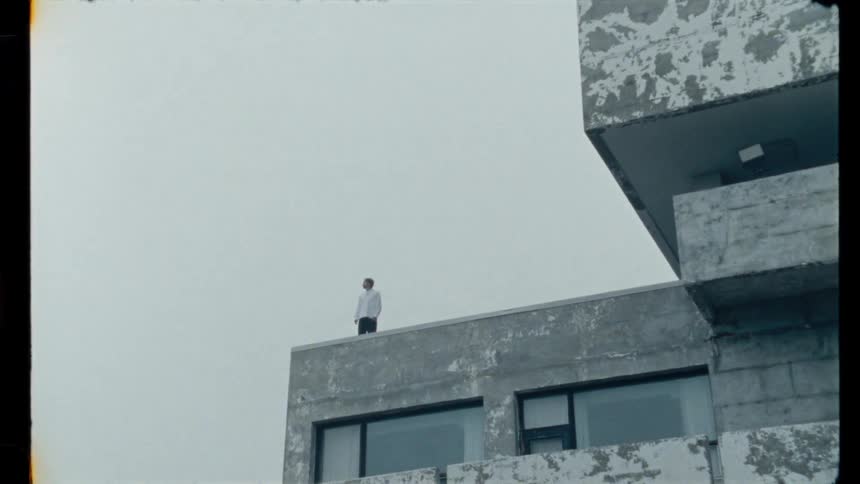 A melancholic man in a white shirt stands alone on the roof of a concrete, brutalist building as the camera zooms out