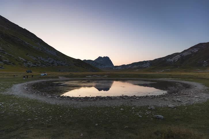 A tranquil timelapse captures the transition from dusk to night over a circular mountain lake, with the peak perfectly reflected in its still waters