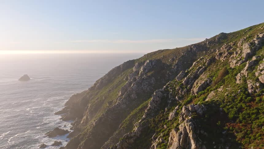 An aerial shot flies toward two hikers standing on a rocky cliff overlooking the vast ocean on a sunny day