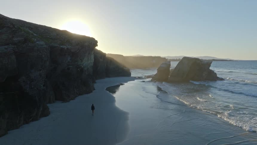 Stunning aerial reveal of a solitary figure walking along a majestic beach with towering rock formations at sunrise
