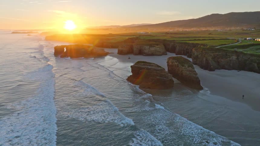 An epic aerial view reveals the stunning Playa de las Catedrales at sunrise, with golden light hitting the natural arches and waves washing ashore
