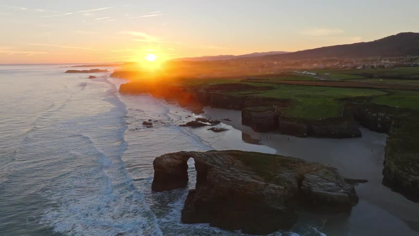 A stunning aerial view pulls back to reveal a magnificent natural rock arch on a sandy beach as the golden sun sets over the ocean waves