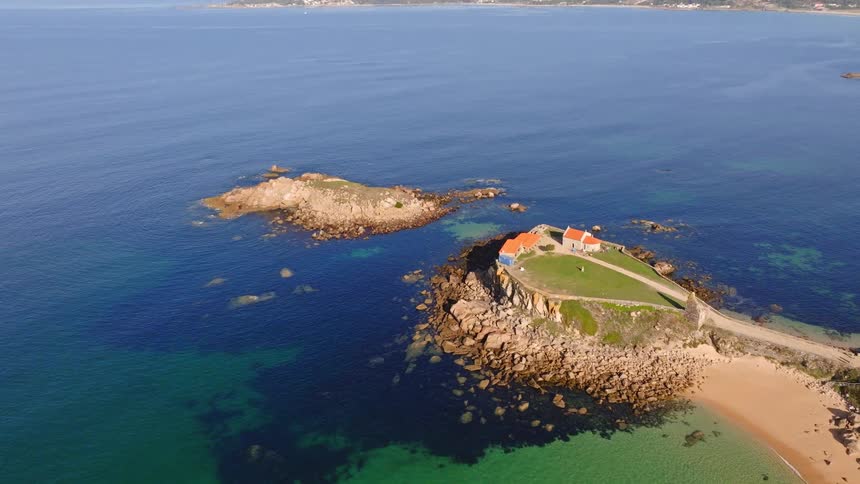 A stunning aerial view tilts up to reveal a picturesque rocky peninsula with a hermitage, a sandy beach, and the expansive blue ocean on a sunny day