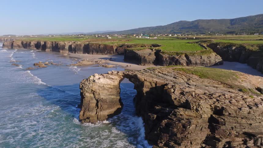 A stunning aerial view pulls back to reveal the famous natural rock arch at Cathedrals Beach, Spain, as waves crash along the picturesque coastline