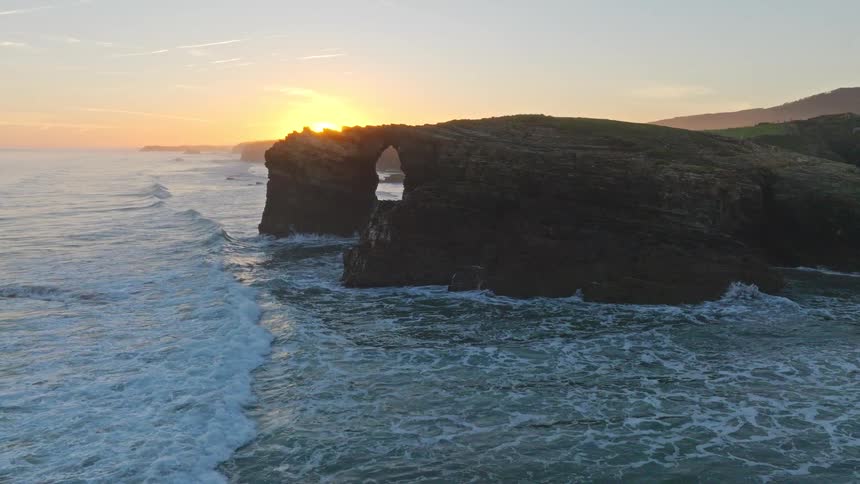An aerial view pulls back revealing the sun rising over the massive sea arches of Cathedrals Beach in Spain as waves crash against the cliffs