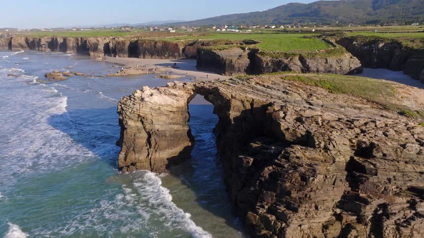 A stunning aerial view flies over the ocean towards the magnificent natural sea arches of Cathedral Beach in Galicia, Spain