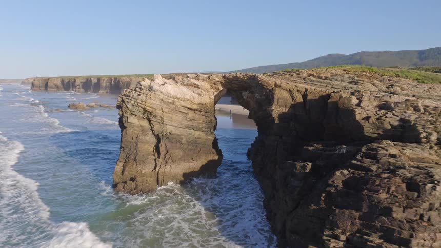 A majestic natural rock arch is revealed on a sunlit coastline as the camera pulls back over the crashing ocean waves