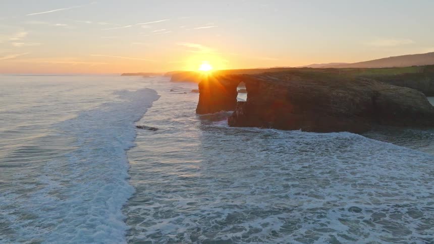 An incredible aerial shot captures the golden sunset over the ocean as waves crash against a majestic sea arch
