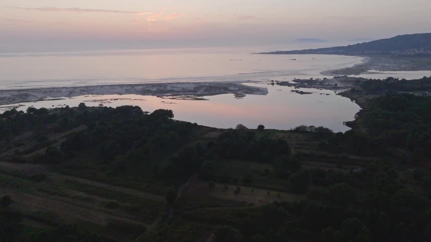 A stunning aerial pullback shot reveals a tranquil coastal lagoon separated from the ocean by a sandbar, surrounded by a lush forest at dusk