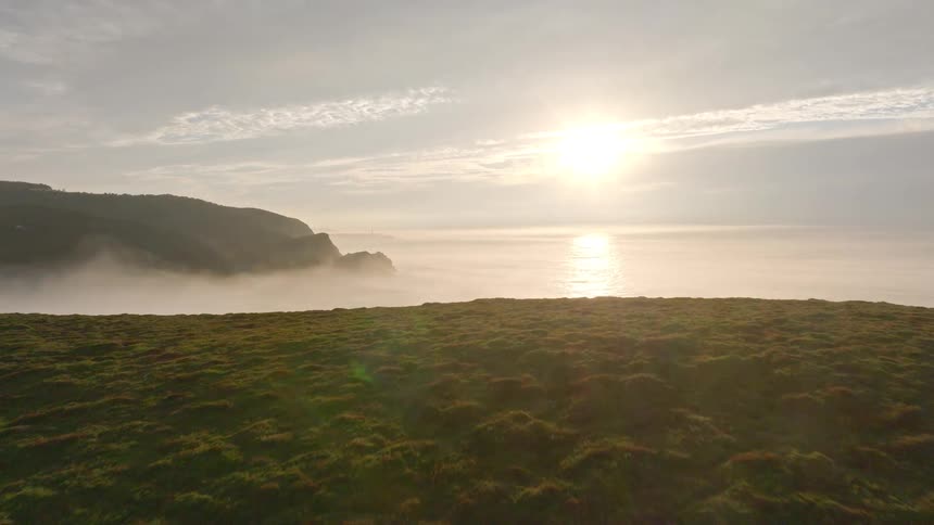 Aerial view pushes past a person standing silhouetted on a camper van at sunrise to reveal a stunning foggy coastline with crashing waves