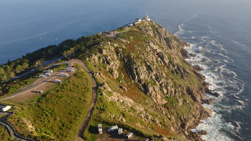 Aerial footage flies towards the stunning Finisterre Lighthouse on a rugged coastal headland, where campers enjoy breathtaking ocean views
