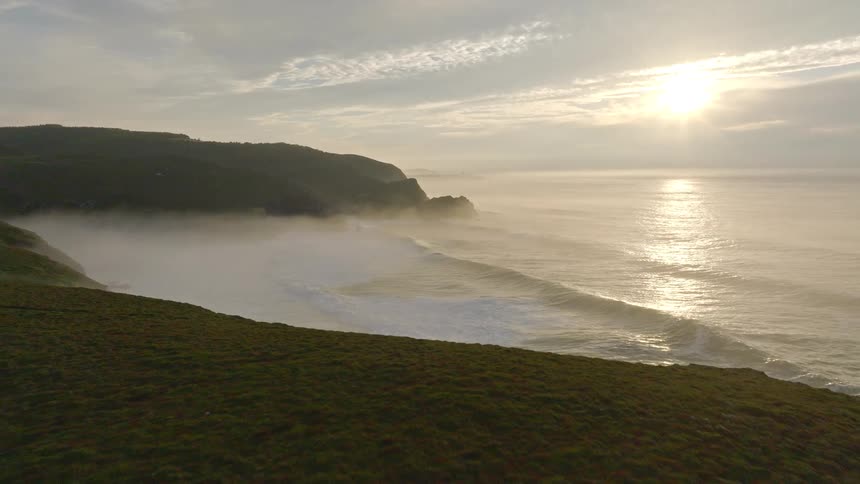 A stunning aerial view pulls back from a misty ocean sunrise to reveal a couple enjoying the view from their camper van on a coastal cliff