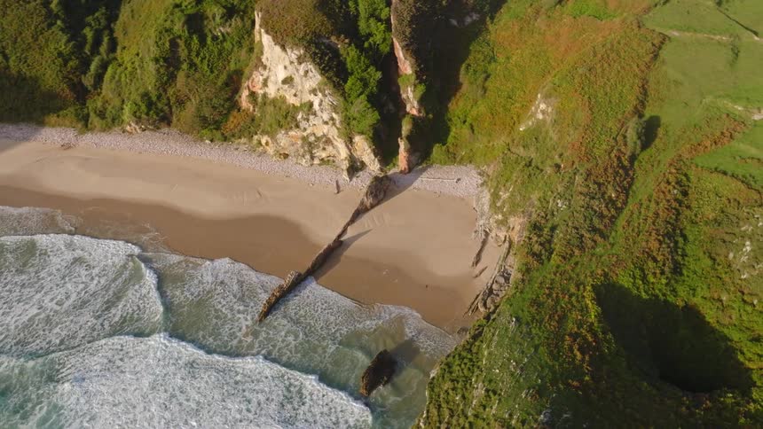A stunning aerial view tilts down over a secluded beach cove, where gentle waves meet golden sand surrounded by verdant cliffs