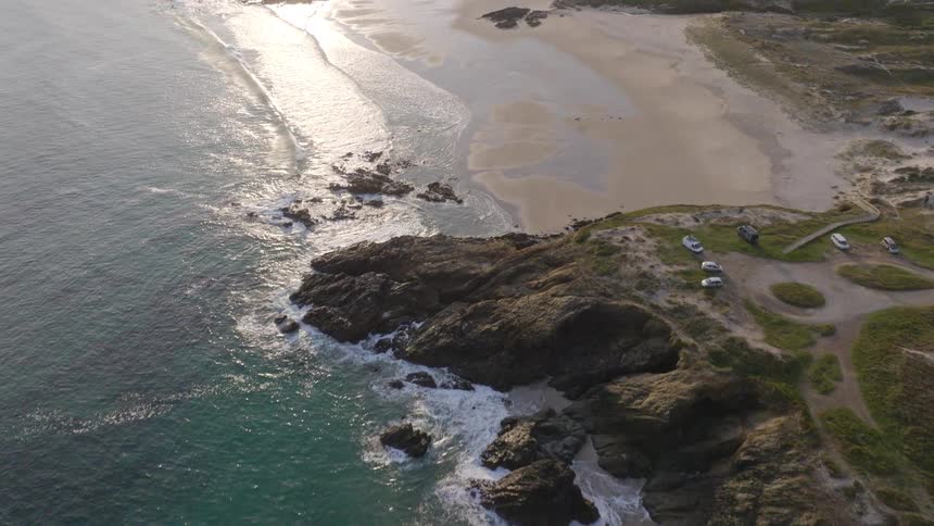 An aerial drone flies along a serene and rugged coastline, revealing a pristine sandy beach and turquoise waves glistening in the warm sunlight