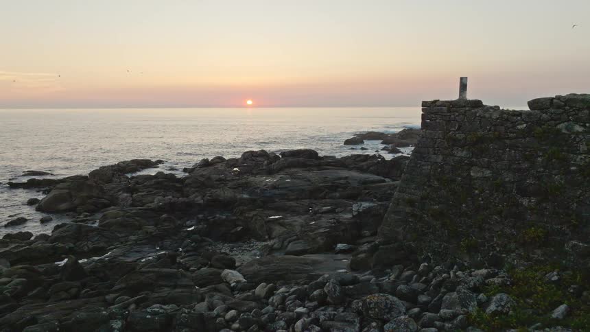 A serene aerial view flies past an ancient stone fort ruin towards the rocky coastline as the sun sets over the ocean
