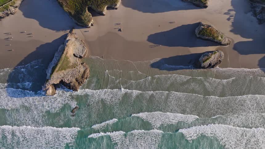 An aerial top-down view shows gentle ocean waves rolling onto a sandy shore with large rock formations and people enjoying a walk on the beach