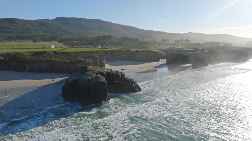 A majestic aerial orbit reveals the stunning sea arches and cliffs of Playa de las Catedrales as tourists enjoy the sunlit beach
