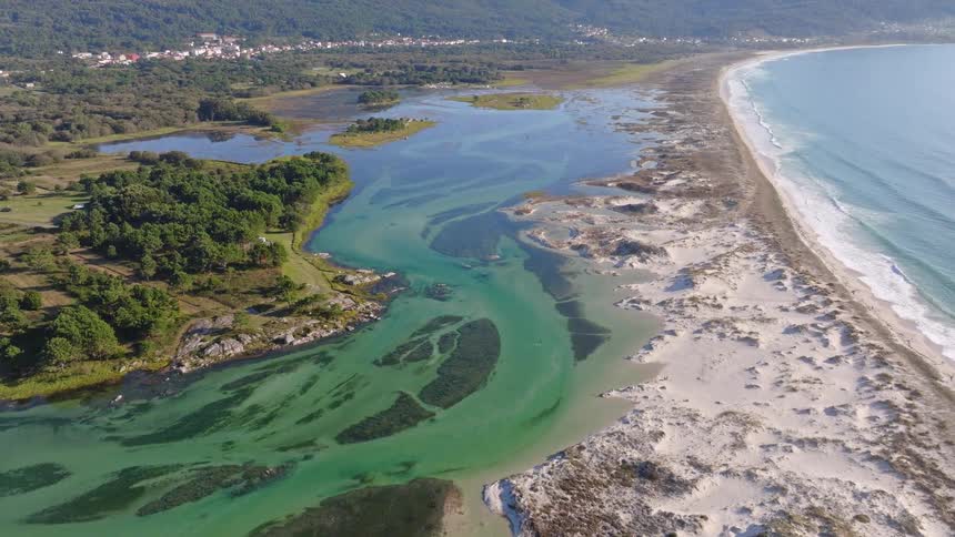 An expansive aerial drone shot slowly flies over a pristine white sand beach and dunes, revealing a beautiful turquoise estuary meeting the blue ocean