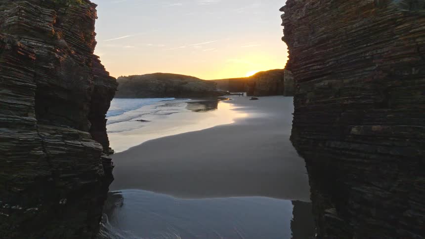 An aerial drone flies between large sea stacks on a tranquil beach, revealing a stunning golden sunset over the ocean