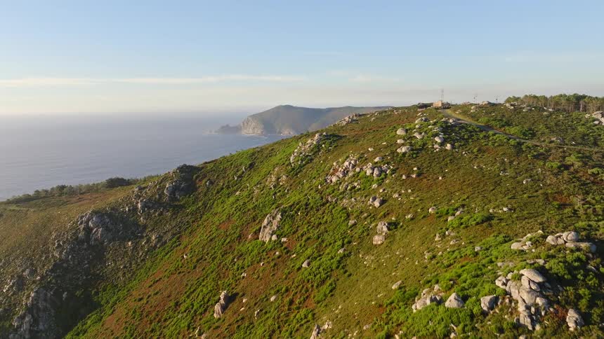 A stunning aerial shot ascends over a rugged green mountain to reveal a beautiful coastal landscape and the vast ocean