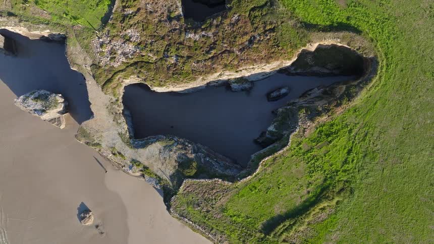 An awe-inspiring top-down aerial shot ascends from a shadowy, hidden cove to reveal a magnificent beach and dramatic coastal cliffs