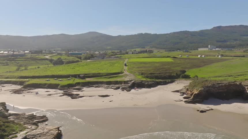 Aerial view reveals a camper van and two people on a rural road leading to a beautiful, hidden beach cove with crashing waves