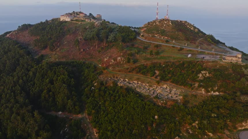 A slow aerial pullback reveals the ancient circular ruins of a Celtic settlement on a coastal mountain in Spain