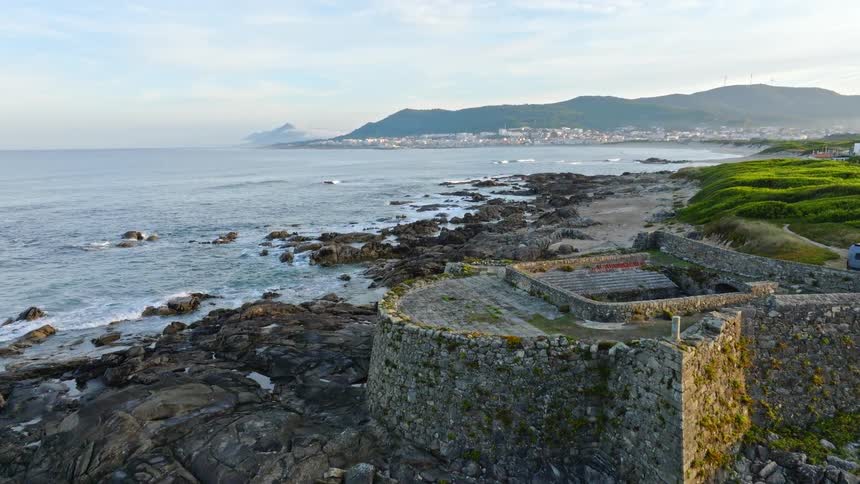An aerial view flies over the historic ruins of a coastal fort with a scenic view of the ocean and a distant town at sunrise