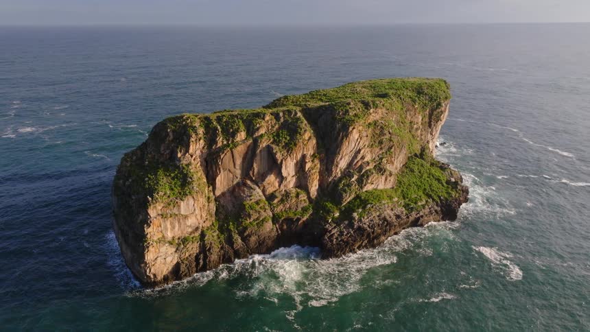 A serene aerial shot circles a majestic, green-topped rocky island standing alone in the vast ocean as golden light hits its cliffs