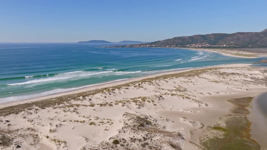 A stunning aerial view flies over a coastal river and sand dunes, revealing a beautiful beach with turquoise waves and a distant mountain landscape