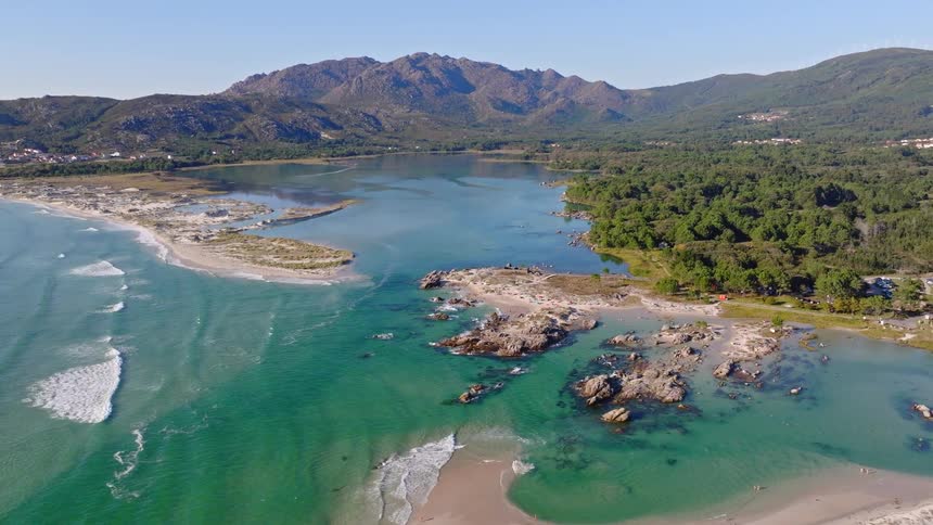 A breathtaking aerial view pulling back to reveal a stunning coastal landscape where a turquoise ocean meets a calm lagoon, bordered by sandy beaches, forests, and mountains