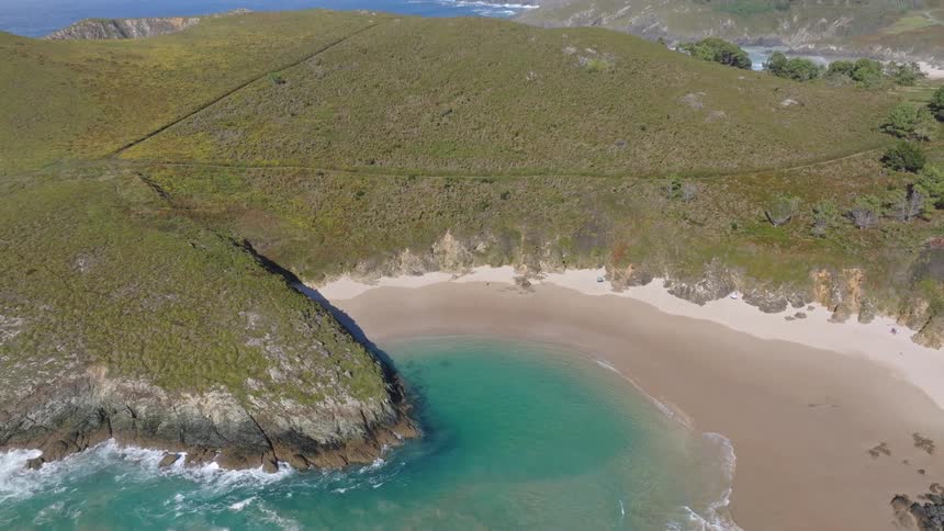 A stunning top-down aerial shot slowly zooms out from a pristine, secluded beach cove with clear turquoise water, revealing a magnificent coastal landscape