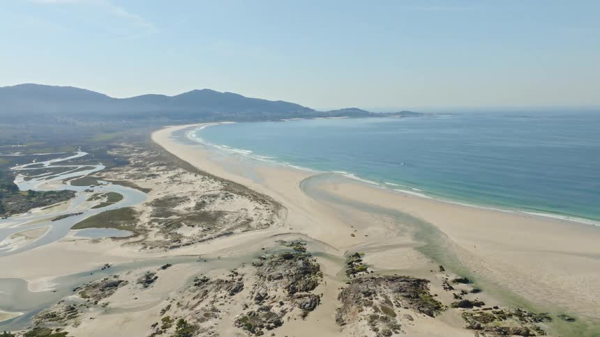 A serene aerial pullback shot reveals a vast sandy coastline where a winding estuary meets the tranquil blue ocean