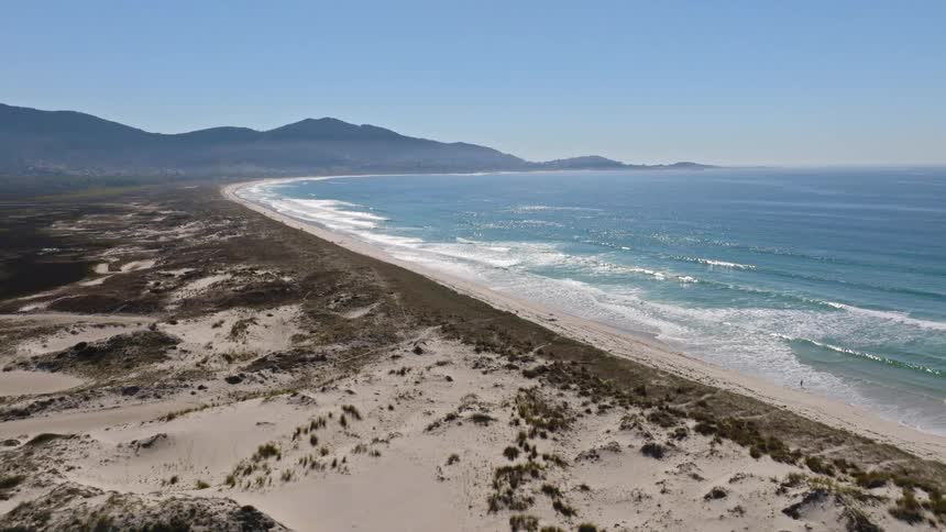 A serene aerial shot reveals a vast coastline with sandy dunes, a long, beautiful beach, and gentle ocean waves under a clear blue sky