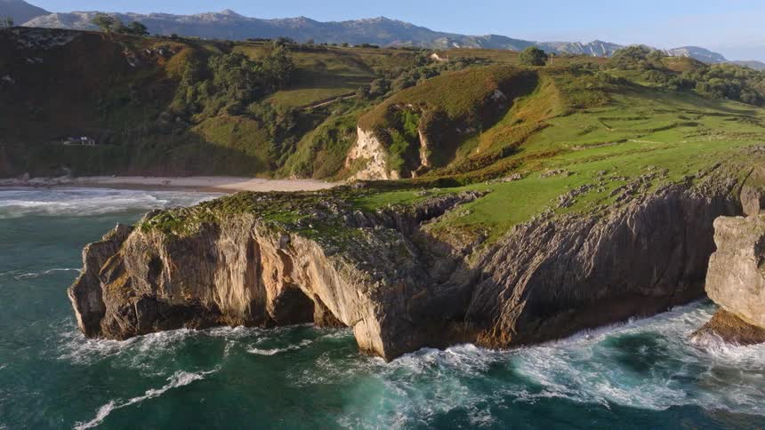 A majestic aerial view captures the lush green cliffs and rugged coastline with a secluded sandy cove as the turquoise ocean waves crash against the shore