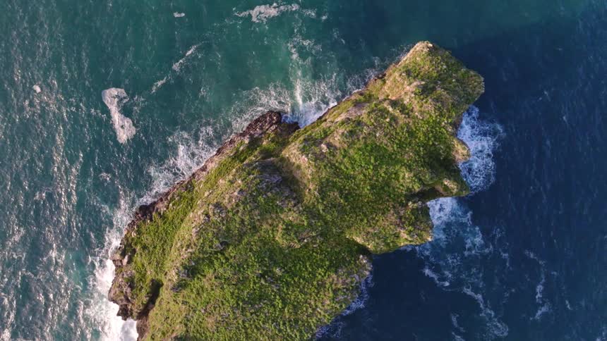 A top-down aerial view slowly pulls up from a verdant, rocky island as turquoise ocean waves crash against its shores